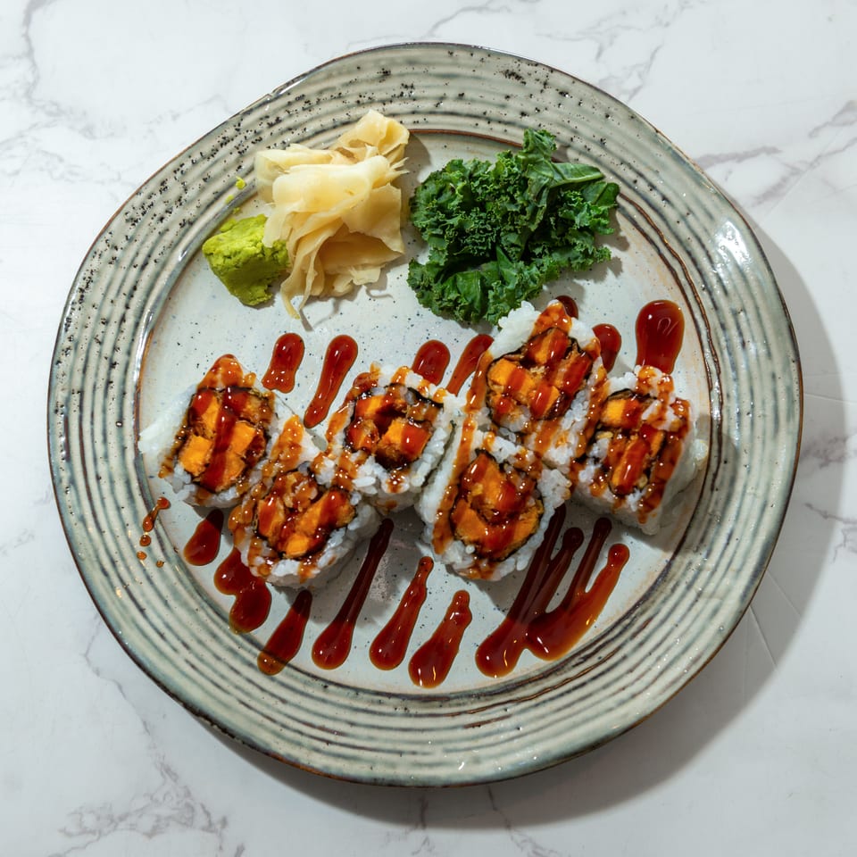 Sushi rolls topped with orange filling and drizzled sauce, served with wasabi, pickled ginger, and kale on a ceramic plate.