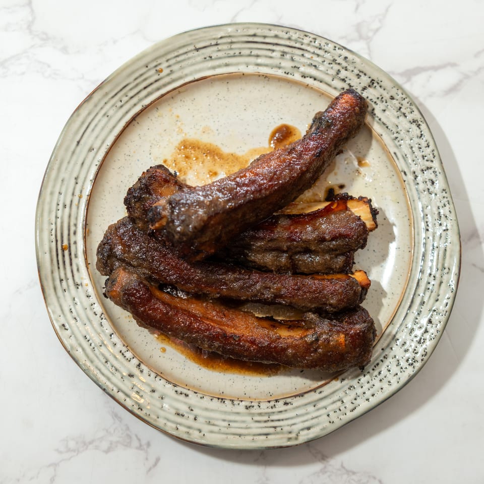 Stack of glazed, roasted beef ribs on a speckled ceramic plate atop a marble tabletop.