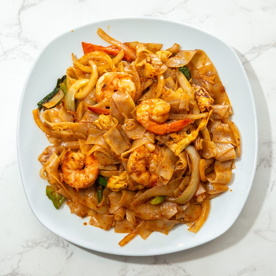 Spicy stir-fried wide rice noodles with shrimp, chicken, onions, peppers, and leafy greens on a white plate.