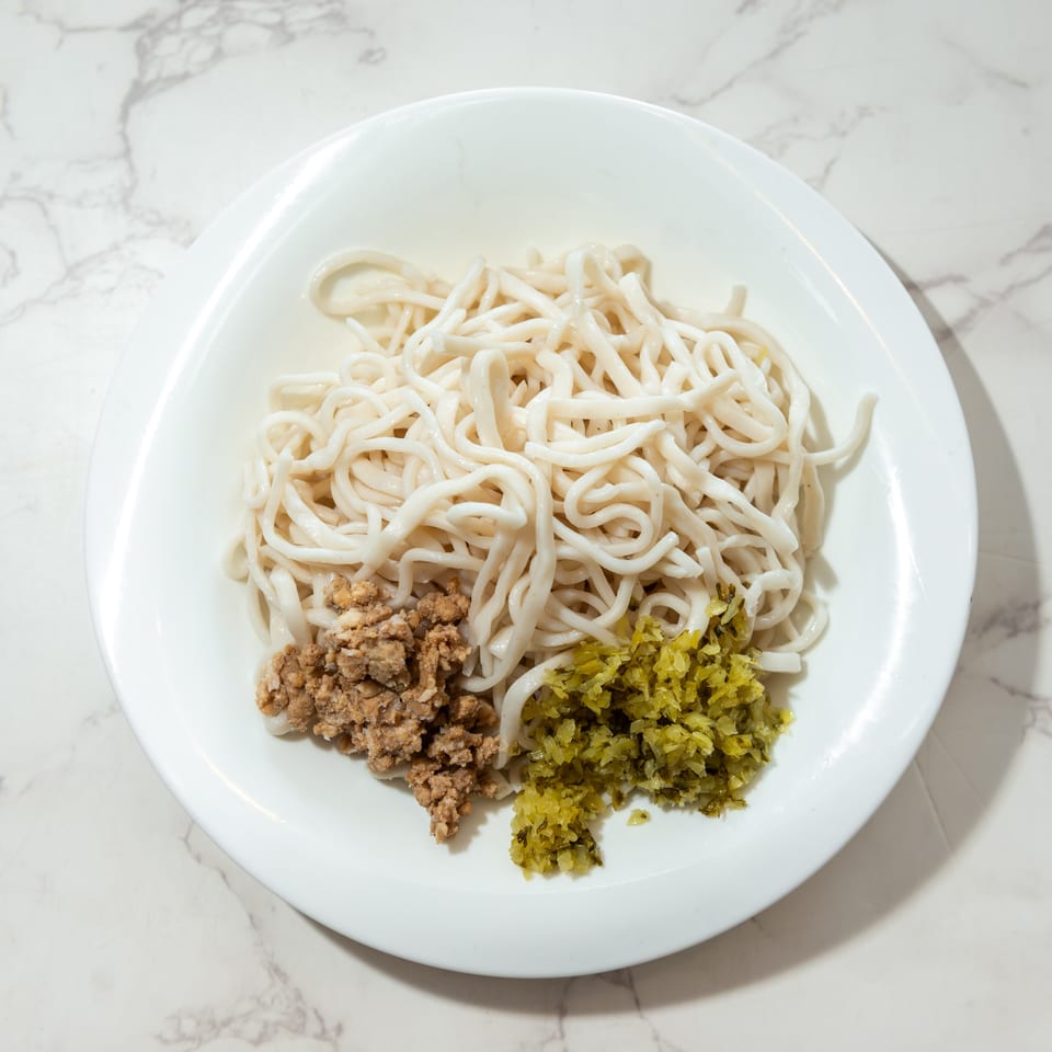 White noodles topped with crumbled meat and chopped green vegetables on a white plate over marble.