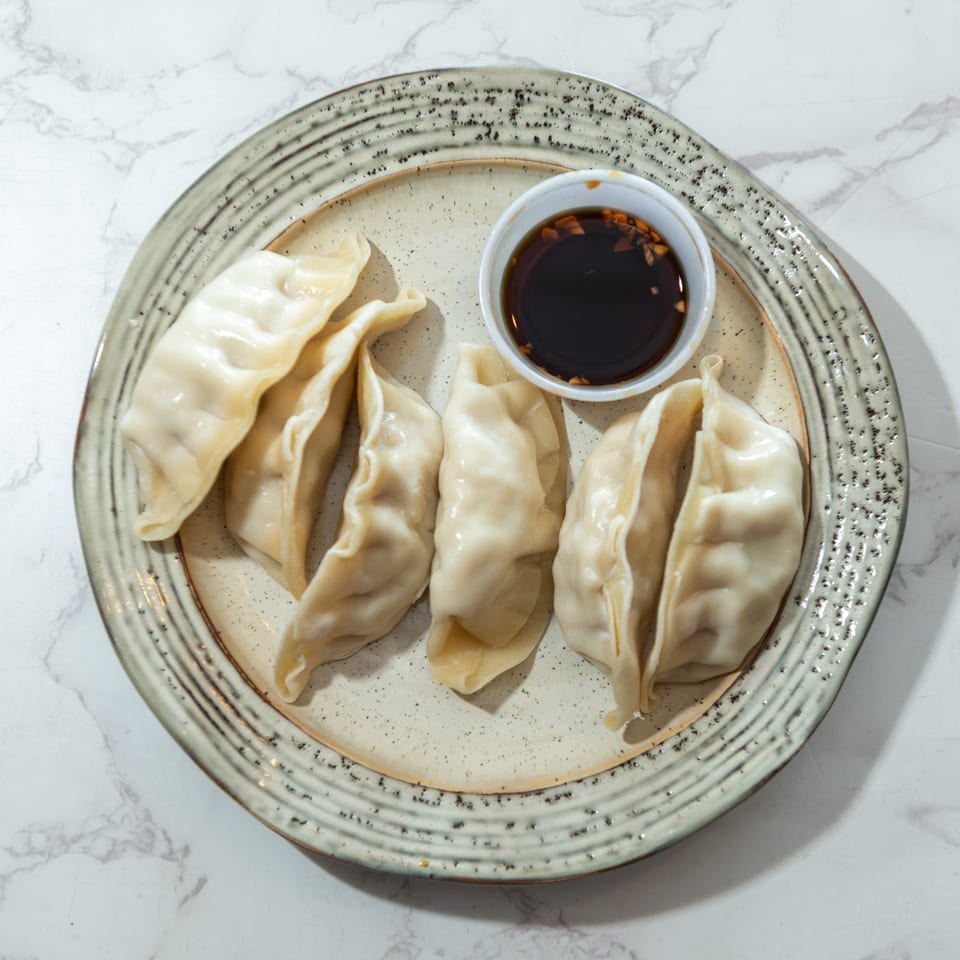 Five steamed dumplings arranged on a ceramic plate with a small bowl of dark dipping sauce.