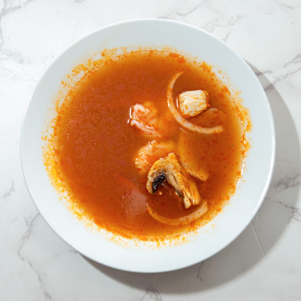 Bowl of red seafood soup with squid rings, shrimp, and mussels on a white plate.