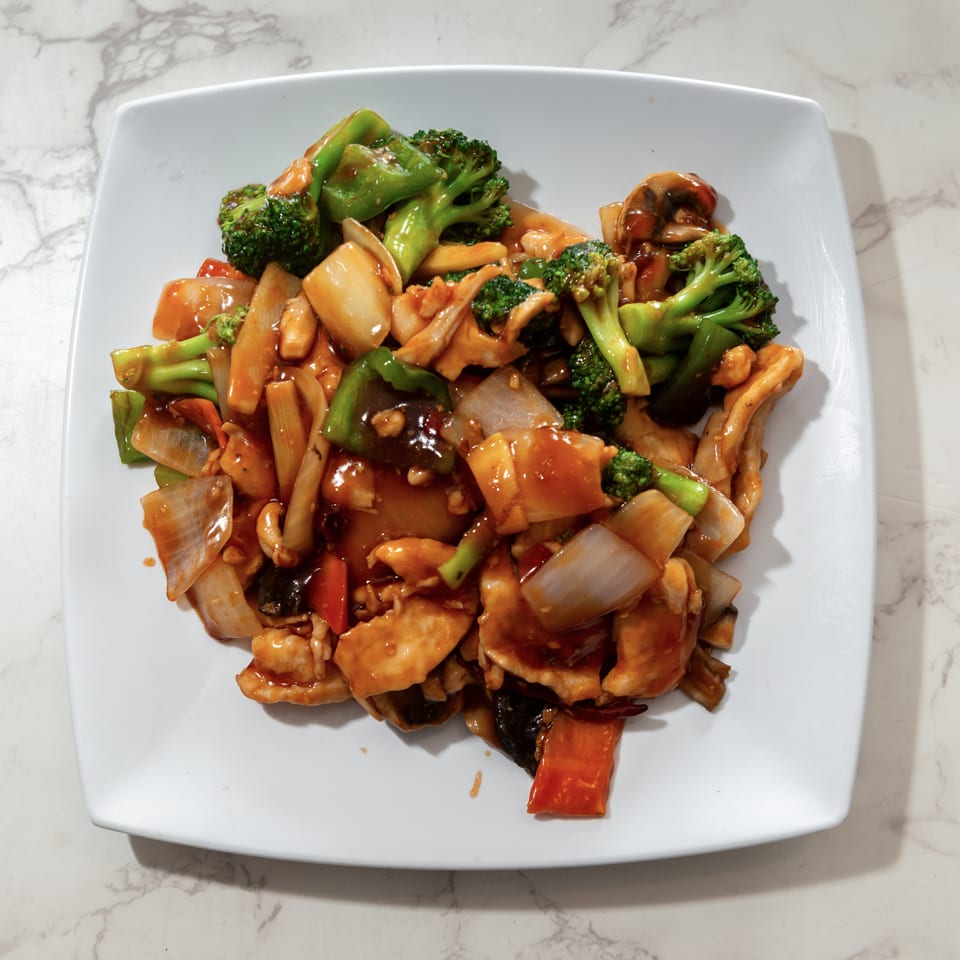 Stir-fried chicken with broccoli, onions, peppers, mushrooms, and carrots in glossy sauce on a white plate.